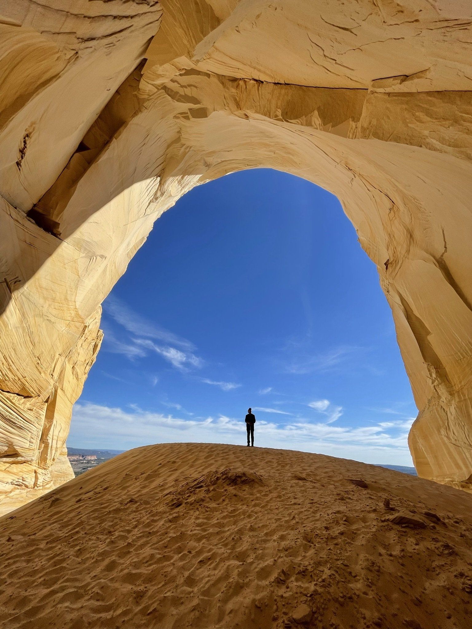 A person stands under a large natural rock arch, looking out at a bright blue sky.