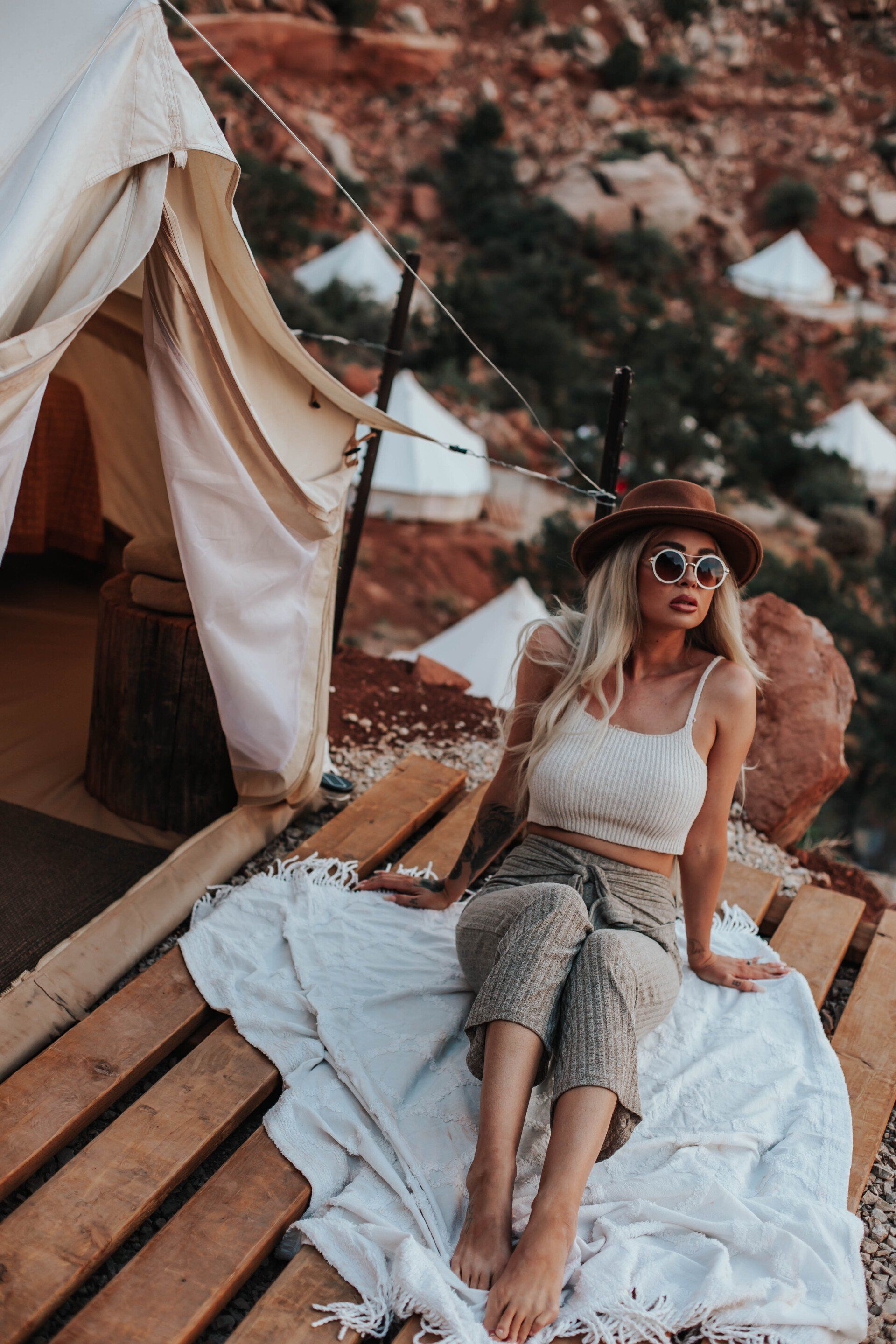 A woman in a hat and sunglasses sits on a blanket outside her glamping tent, enjoying the view.