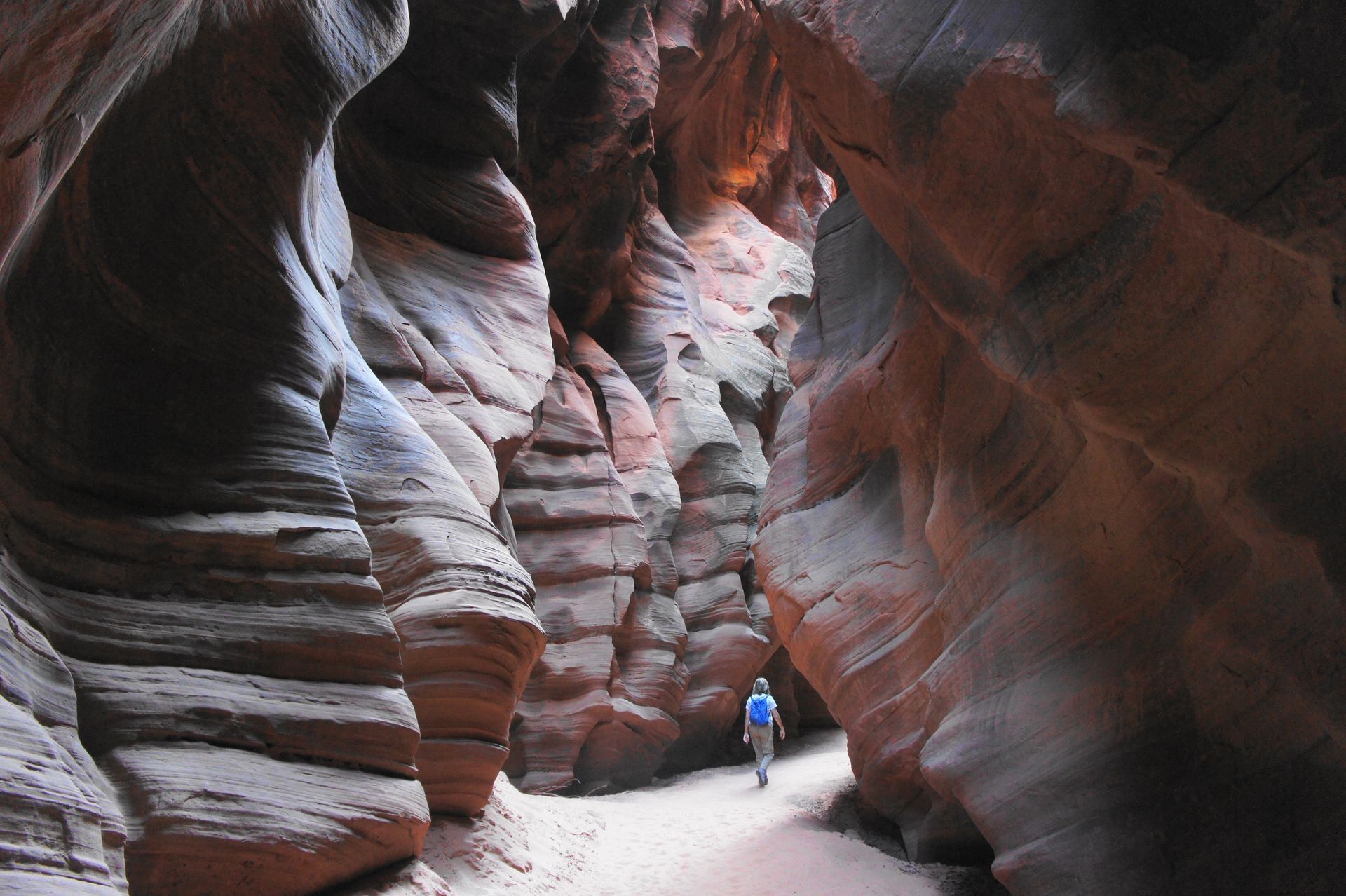 Person walking through a narrow, winding slot canyon with smooth, wavy rock walls.