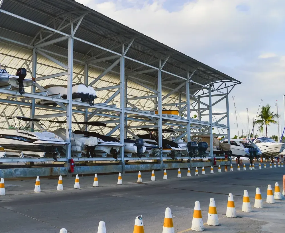 Boats stored on multi-level racks under a metal roof in a marina; orange and white cones line the parking area. — Gympie Southside Mini Storage in Southside, QLD
