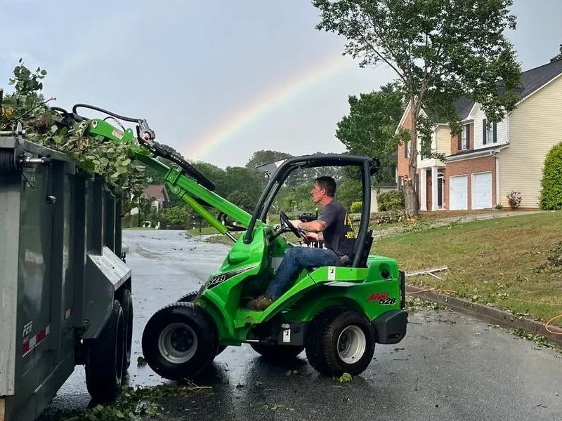 Man operating a green front-end loader, loading debris into a trailer on a street with a rainbow in the background.