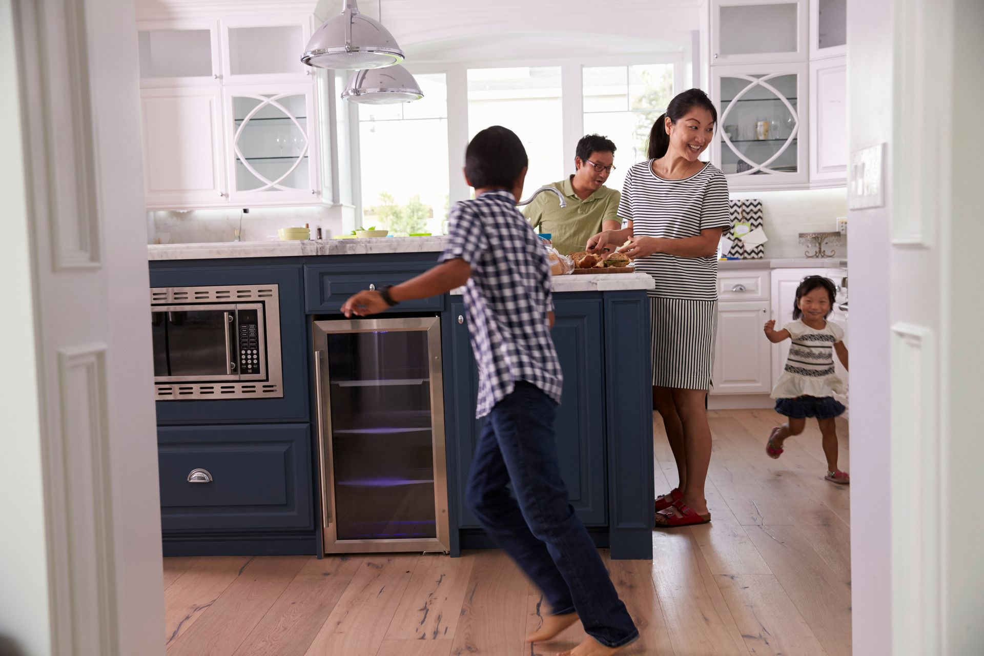 A family moves through a bright kitchen featuring blue cabinets, a built-in microwave, and a glass-front beverage cooler.