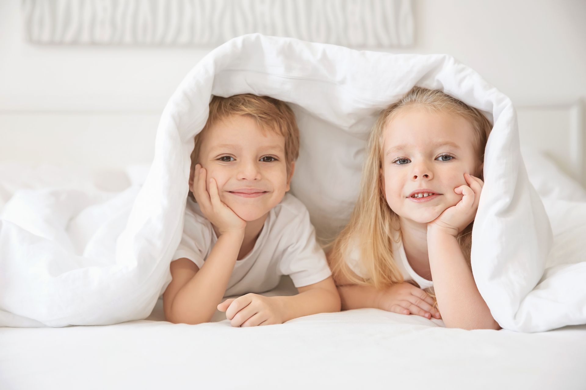 Two children with smiling expressions resting their chins on their hands while nestled together under a white blanket. Two children with smiling expressions resting their chins on their hands while nestled together under a white blanket.