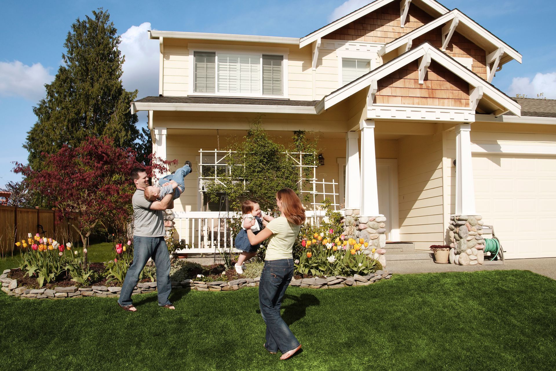 A family plays in the yard in front of a light-yellow two-story house with a green lawn and blooming flowers. A family plays in the yard in front of a light-yellow two-story house with a green lawn and blooming flowers.