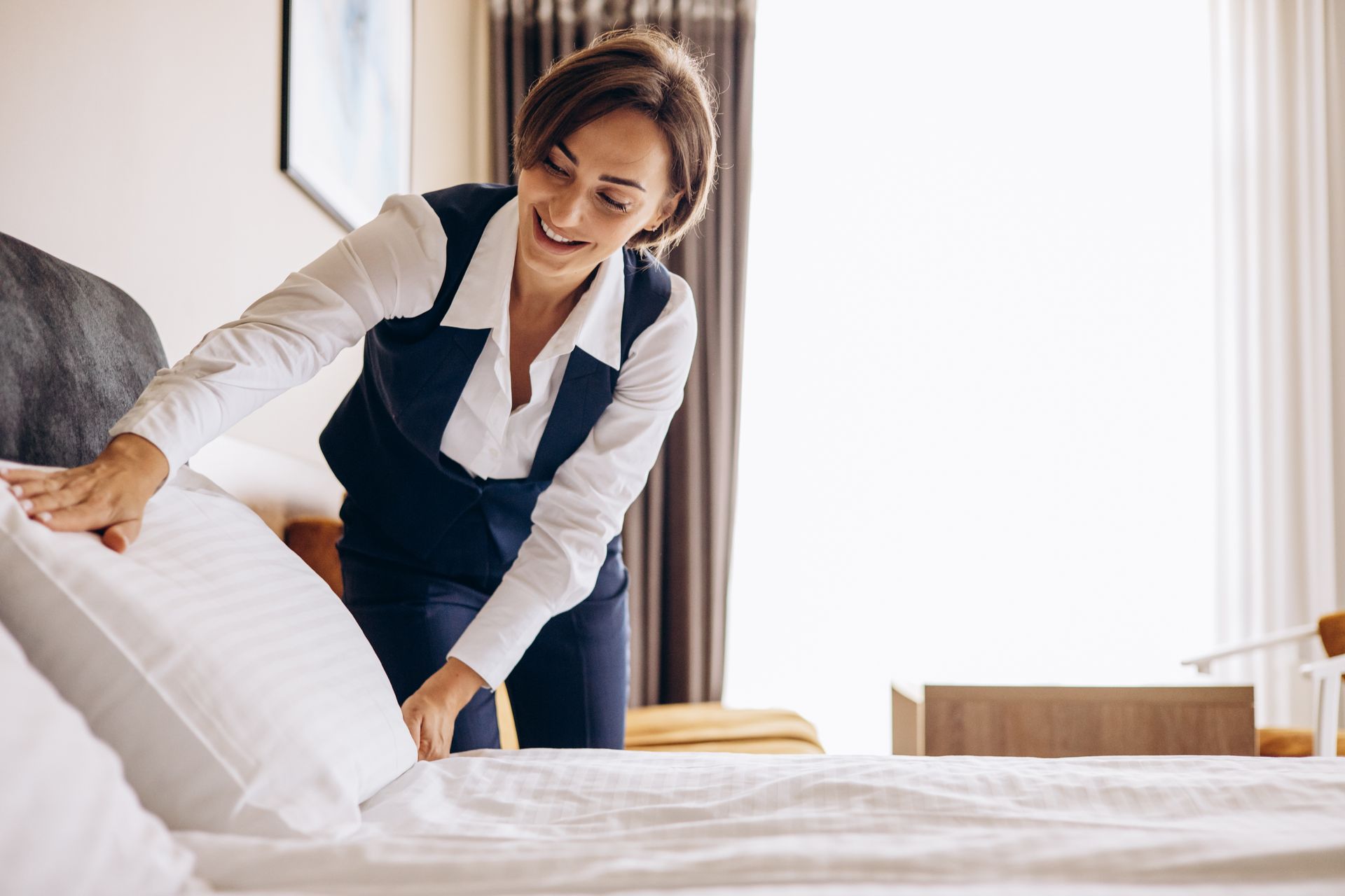 A hotel housekeeper in a white shirt and dark vest smiling while arranging a pillow on a neatly made bed in a guest room. A hotel housekeeper in a white shirt and dark vest smiling while arranging a pillow on a neatly made bed in a guest room.