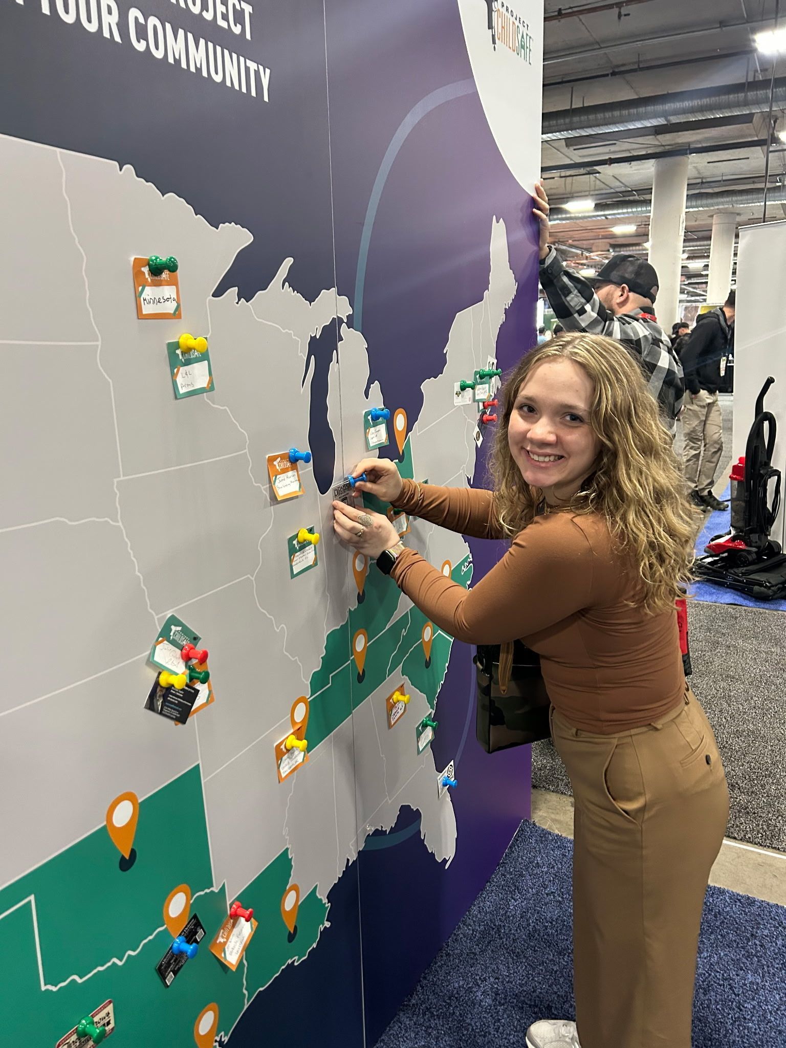 A person with curly hair smiles while placing a pin on a large map of the United States at a display booth.