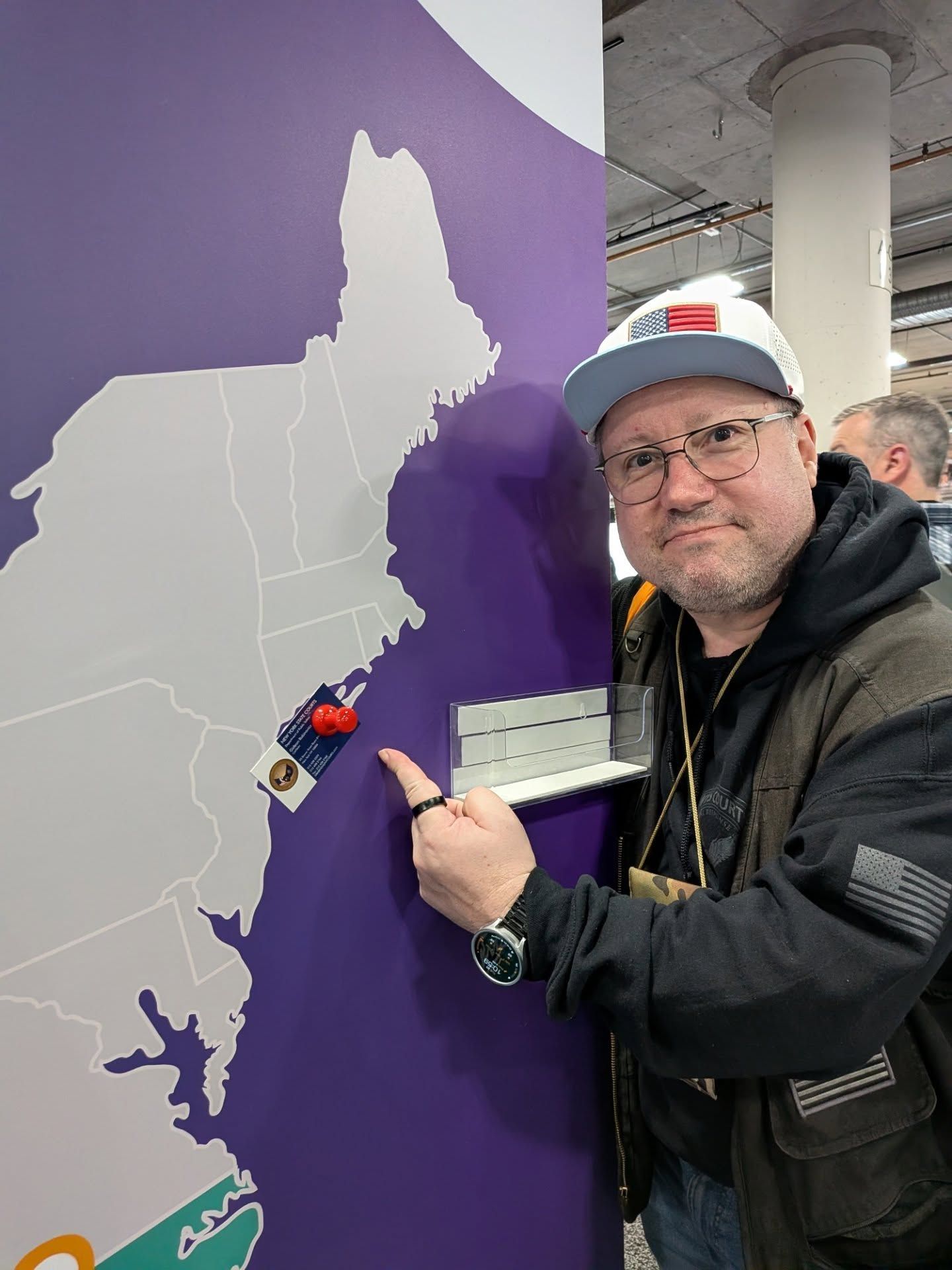 A person in a hat and jacket points to a pin on a wall map of the Northeastern United States in an indoor setting.