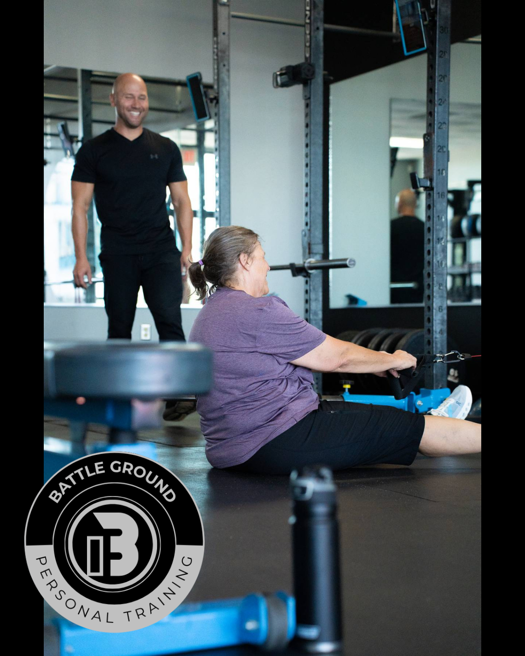 A woman seated on floor exercising with a cable machine; a trainer watches and smiles in gym.