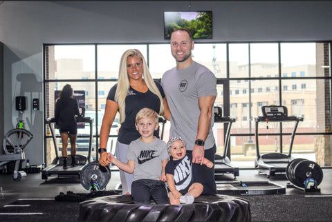 Family of four poses in a gym, two young boys sitting on a tire, woman and man standing.