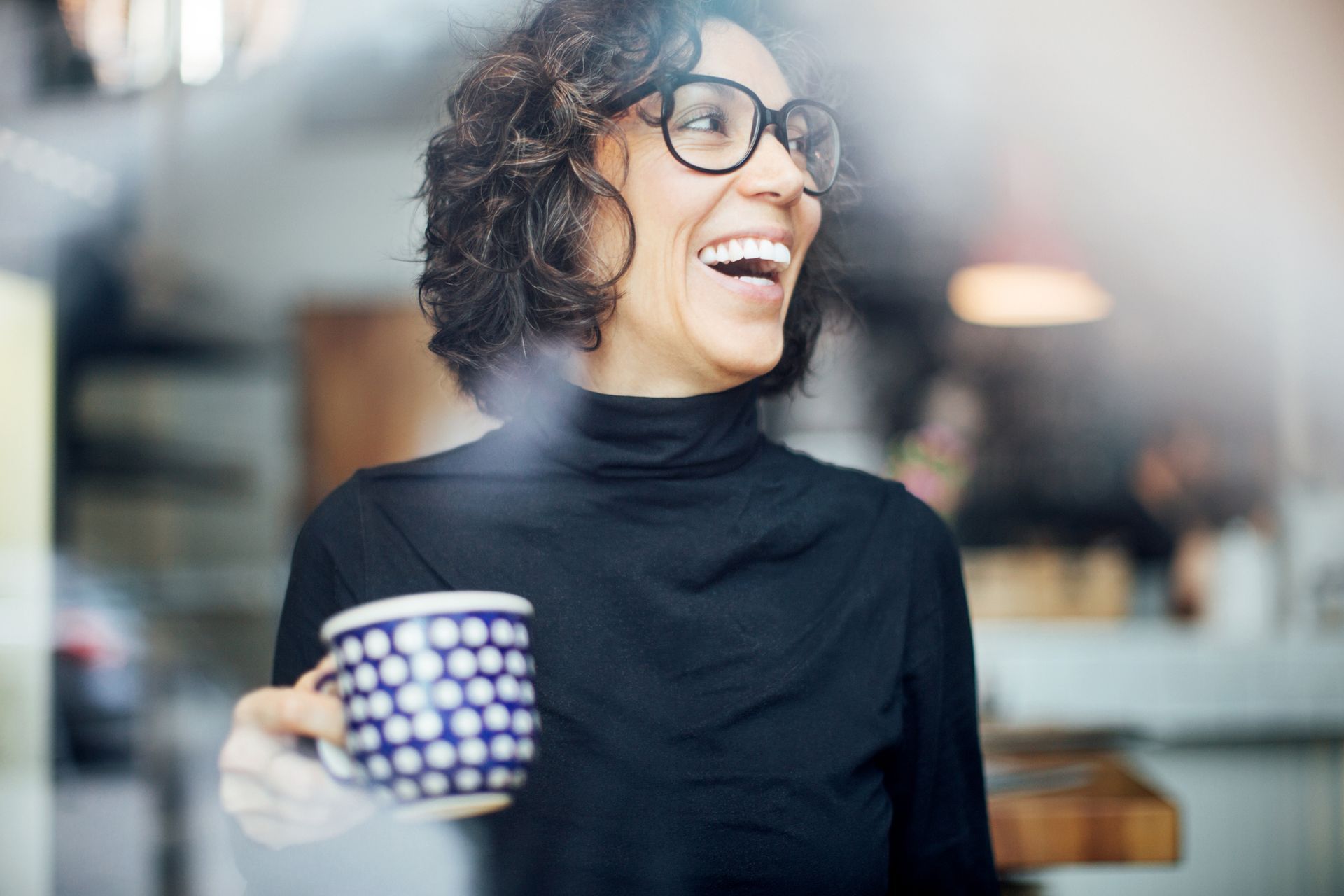 A woman wearing glasses is holding a cup of coffee and smiling.