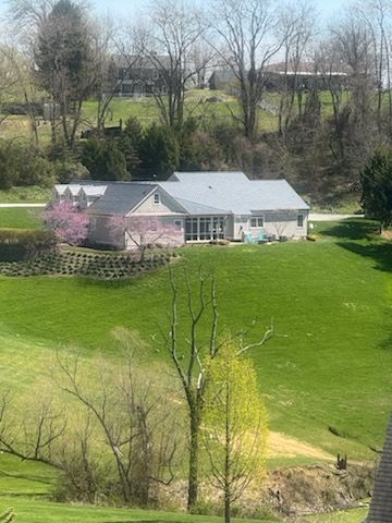 Countryside home with gray roof, pink blossoming tree, and green lawn beside a wooded hillside.