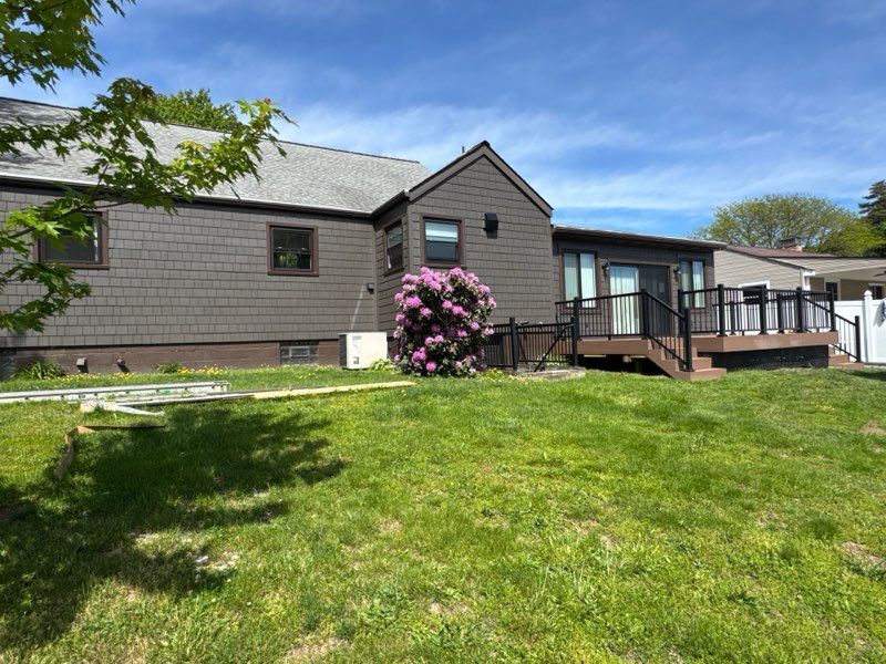 Gray house with a green lawn, pink flowering bush, and wooden deck under a blue sky