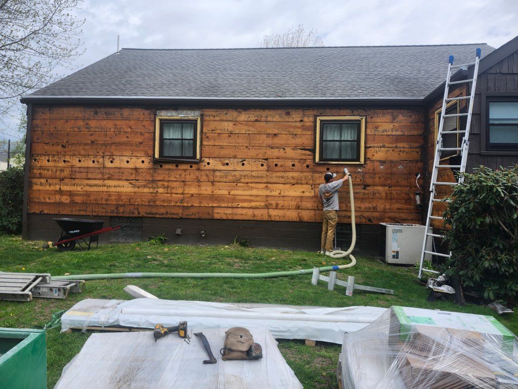 Backyard with a wooden house wall, ladder, and covered work materials on the grass under an overcast sky
