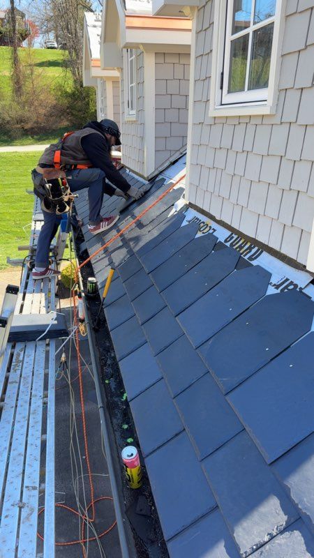 Workers installing dark blue roof shingles on a house beside a window and gutter, with scaffolding below.