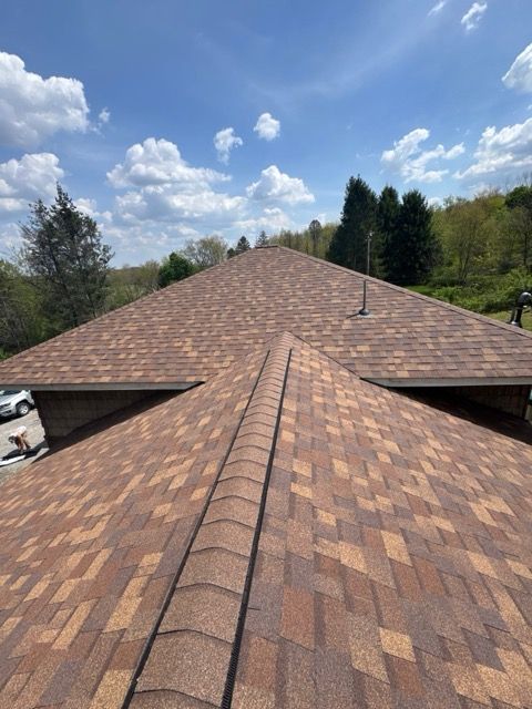 Brown shingle roof with a central ridge line under a partly cloudy blue sky, surrounded by trees