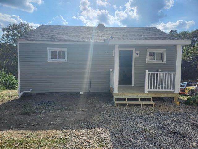Small gray house with a porch, two windows, and dark doorway, gravel driveway.