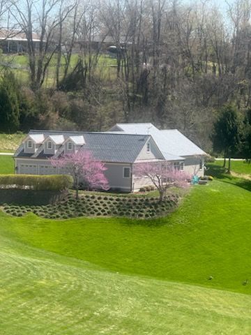 Small house with gray roof by a bright green lawn, pink flowering trees, and a wooded hillside background.