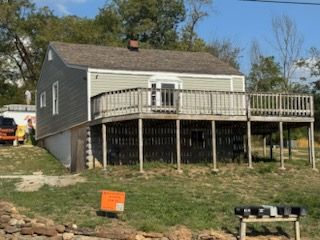 Small house with gray siding, large deck, and brown roof. Situated on a grassy hill, likely rural.