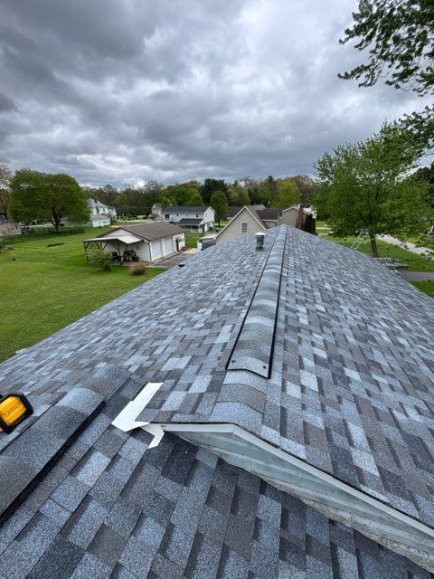 Roofing shingles viewed from a roof ridge under cloudy sky, with neighborhood houses and trees below