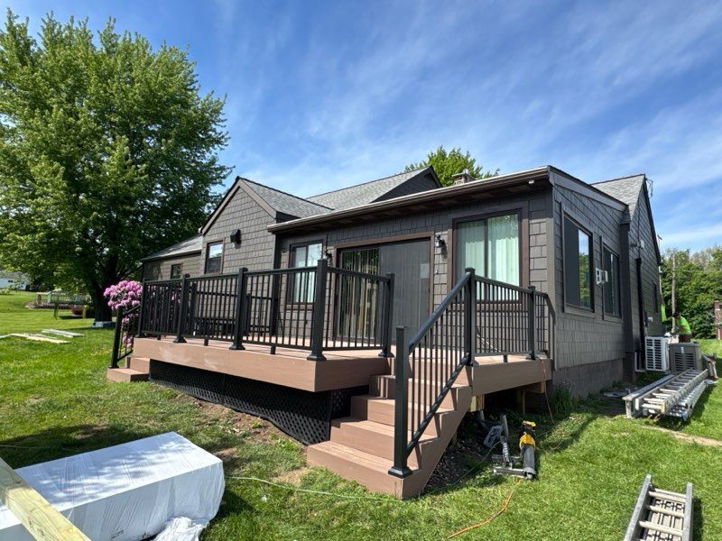 Black house with a raised wooden deck and stairs in a grassy yard under a blue sky