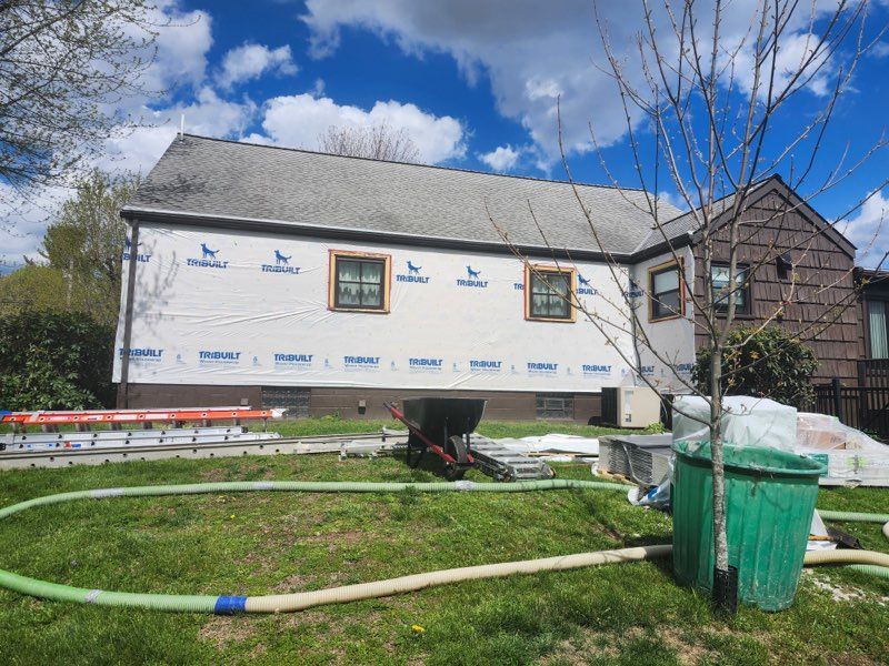 House under construction wrapped in white housewrap, with scaffolding and a green yard hose in front.