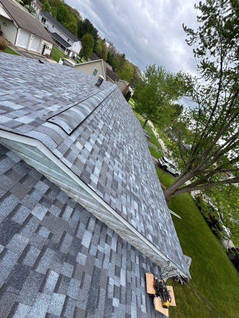 Tilted view of a gray shingled roof with trees and houses beside it under cloudy sky