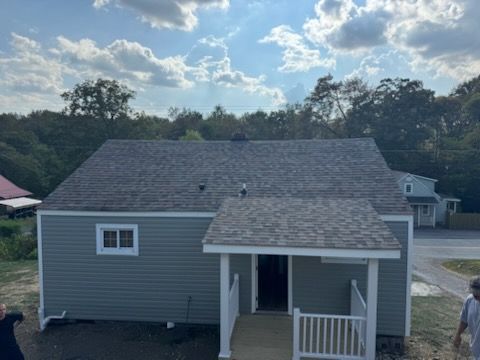 Gray house with a front porch, blue siding, gray roof, and white trim. Blue sky and trees in the background.
