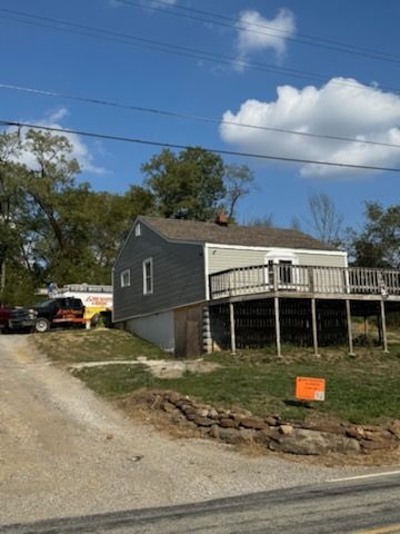 House with gray siding and deck, next to a gravel driveway. A truck is parked to the left.