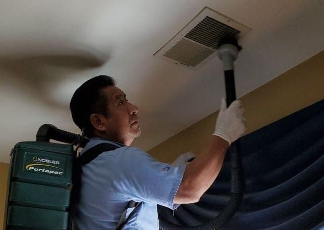 A man is using a backpack vacuum to clean a vent on the ceiling