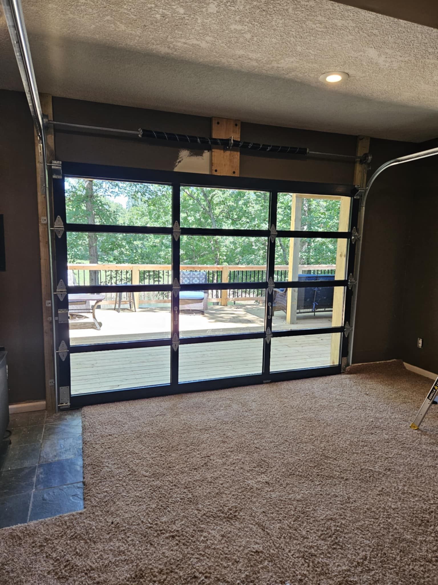 A garage door with a view of a deck and trees is open in a living room.
