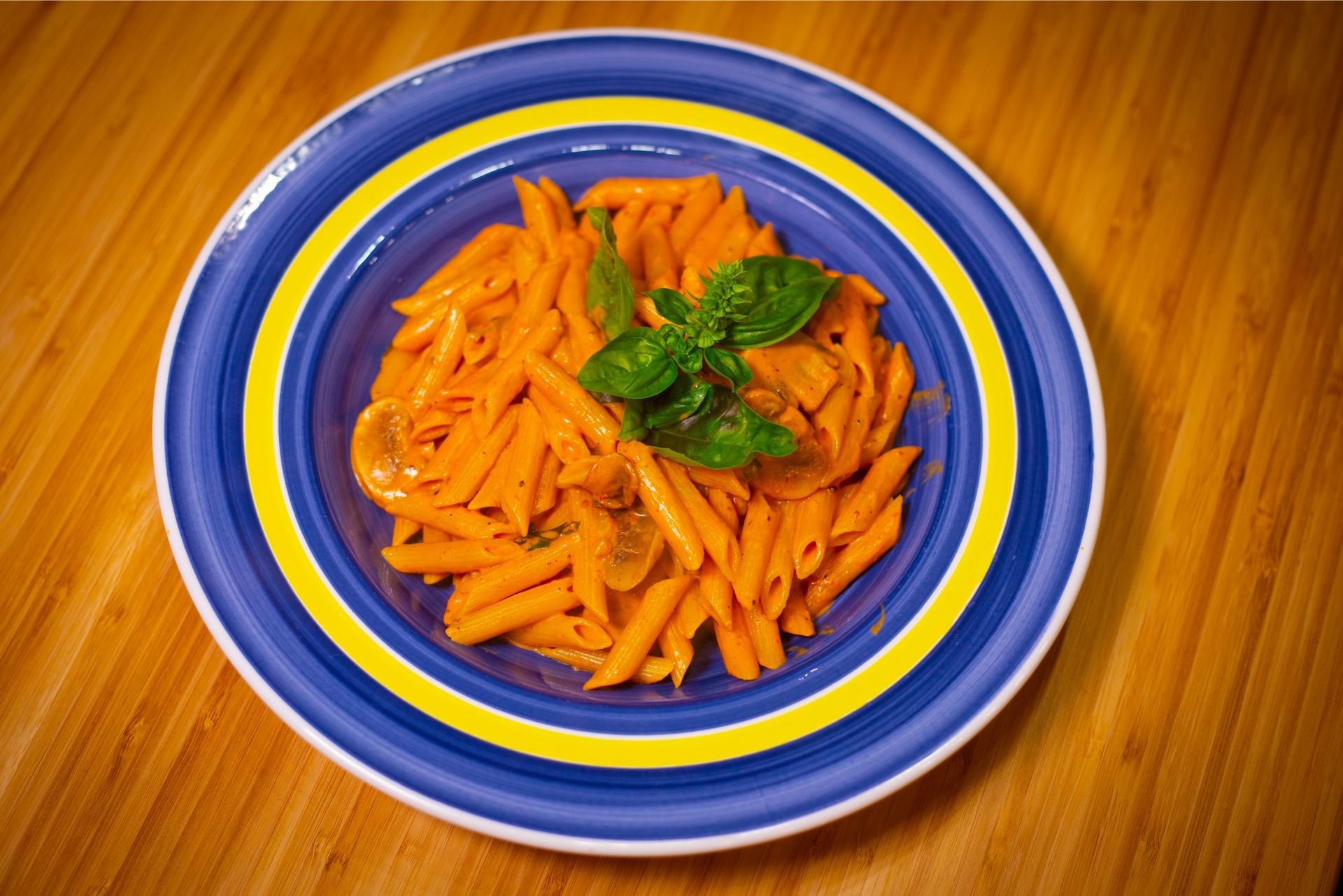 A blue and yellow plate topped with pasta and basil on a wooden table.