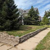 Sidewalk leads past stone wall and grassy hill toward house with trees.