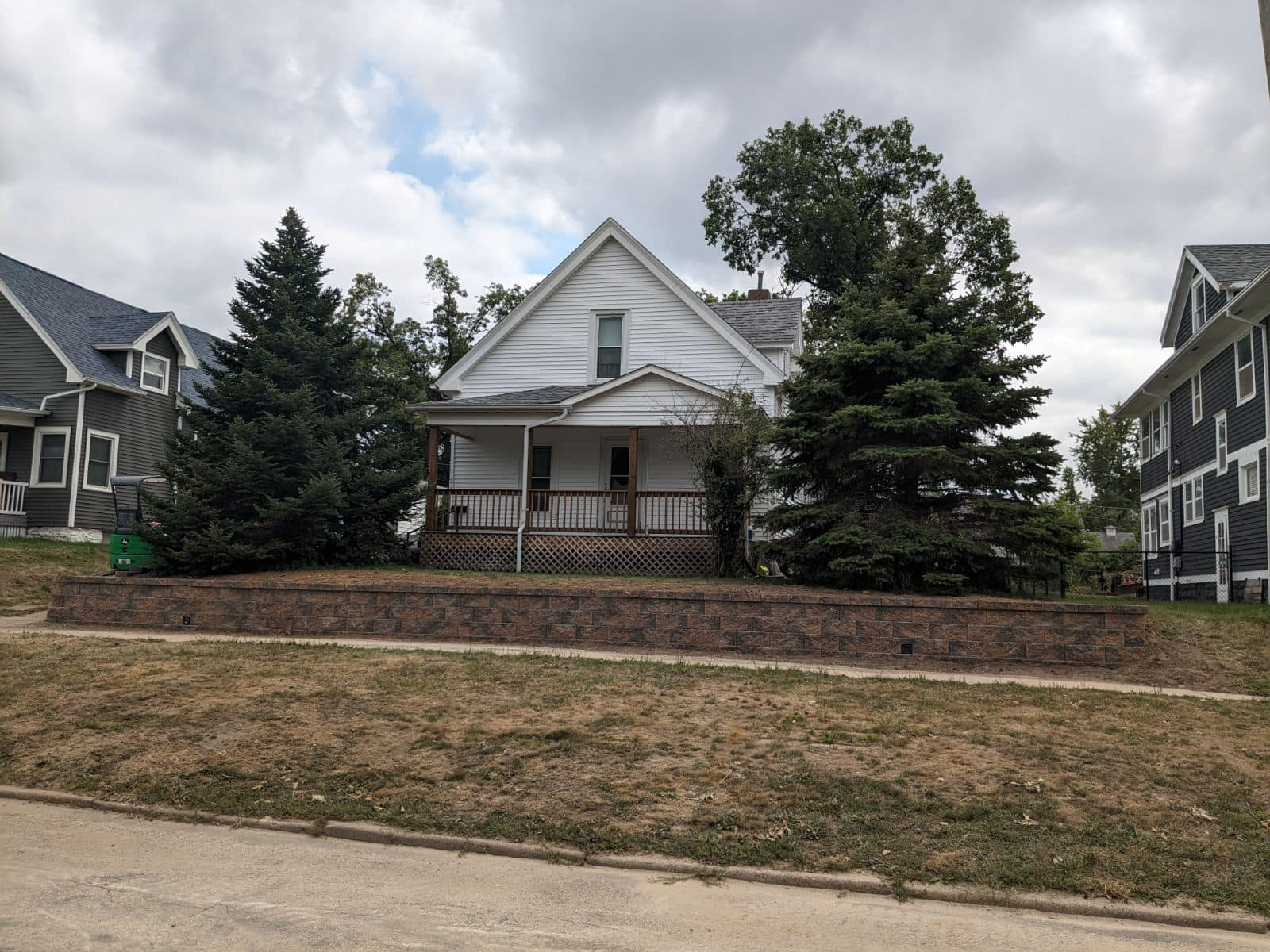 White house with a small porch, flanked by green evergreen trees, against a cloudy sky.