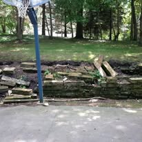 Basketball hoop beside a crumbling stone retaining wall and a concrete surface. Trees in the background.