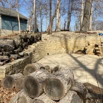 Logs stacked in foreground, concrete patio, stone wall, and trees under a sunny sky.