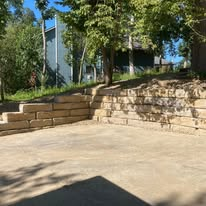 Stone retaining wall next to a paved area, with steps leading up to a grassy slope and trees.