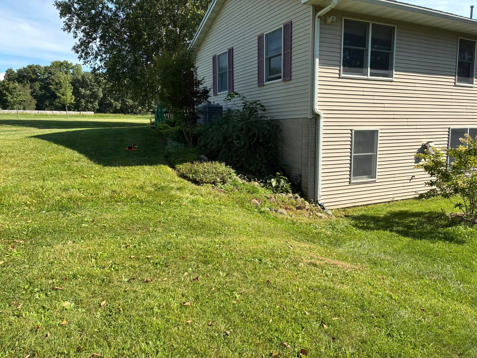 Exterior view of a house with a metal chimney and wooden retaining wall on a sloped yard covered in red mulch and green plants.
