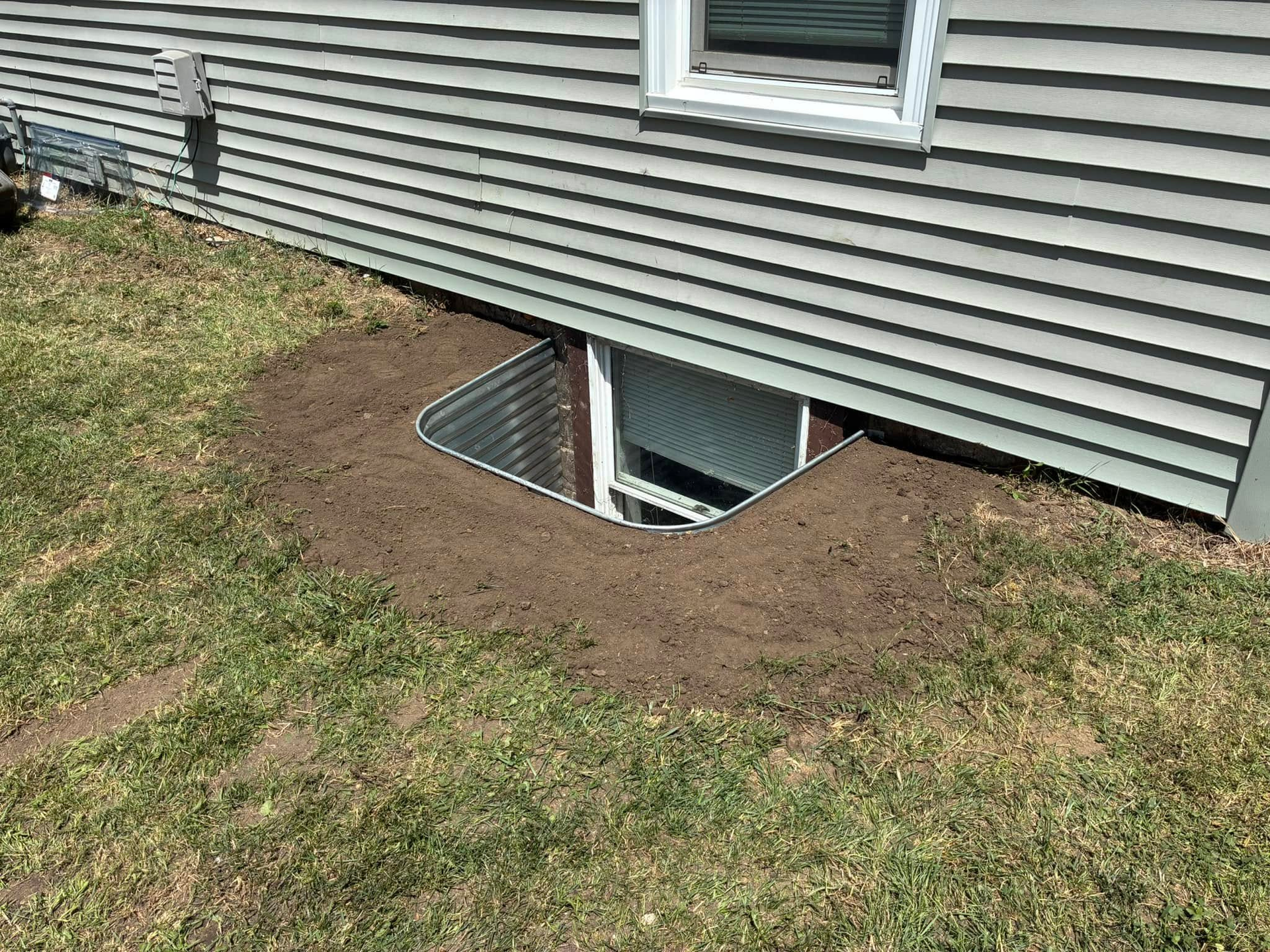 Exterior view of a house with window well surrounded by dirt and grass.