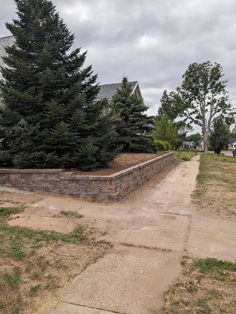 Sidewalk leads past stone wall and grassy hill toward house with trees.