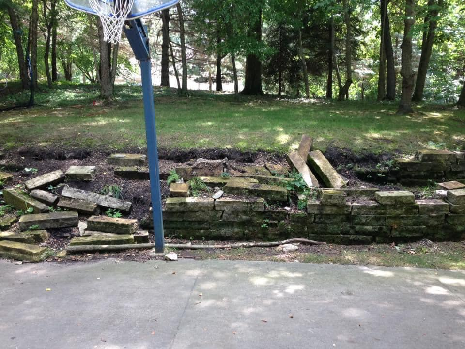 Basketball hoop beside a crumbling stone retaining wall and a concrete surface. Trees in the background.