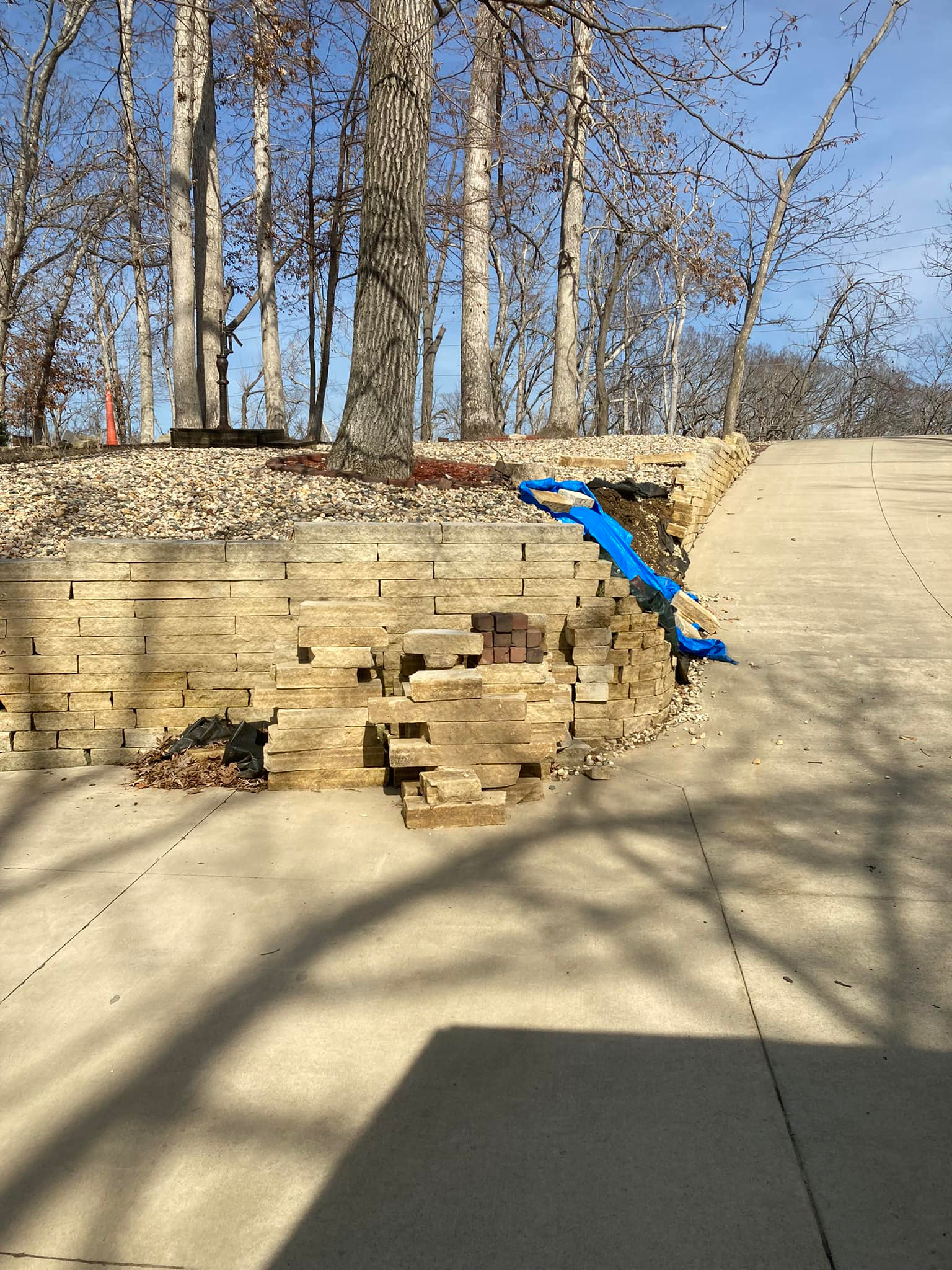 Logs stacked in foreground, concrete patio, stone wall, and trees under a sunny sky.