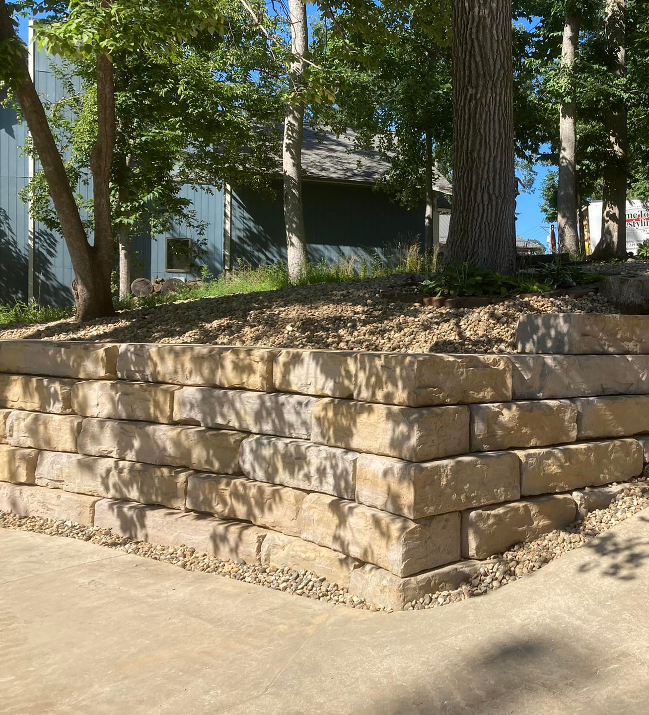 Stone retaining wall next to a paved area, with steps leading up to a grassy slope and trees.