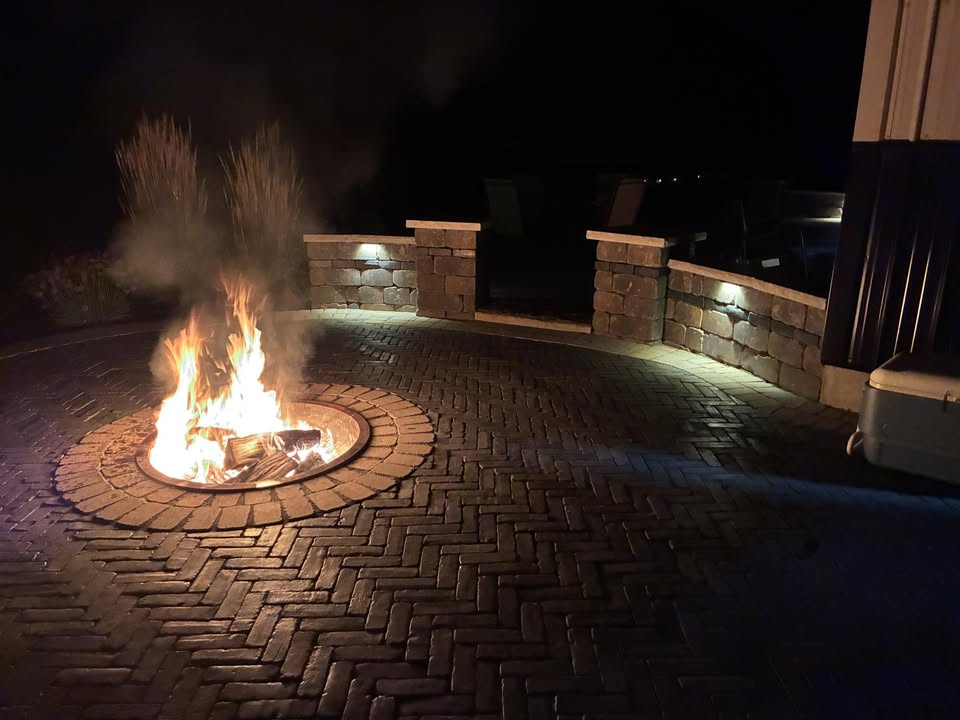 Fire pit ablaze on brick patio at night. Stone wall with lights in background.