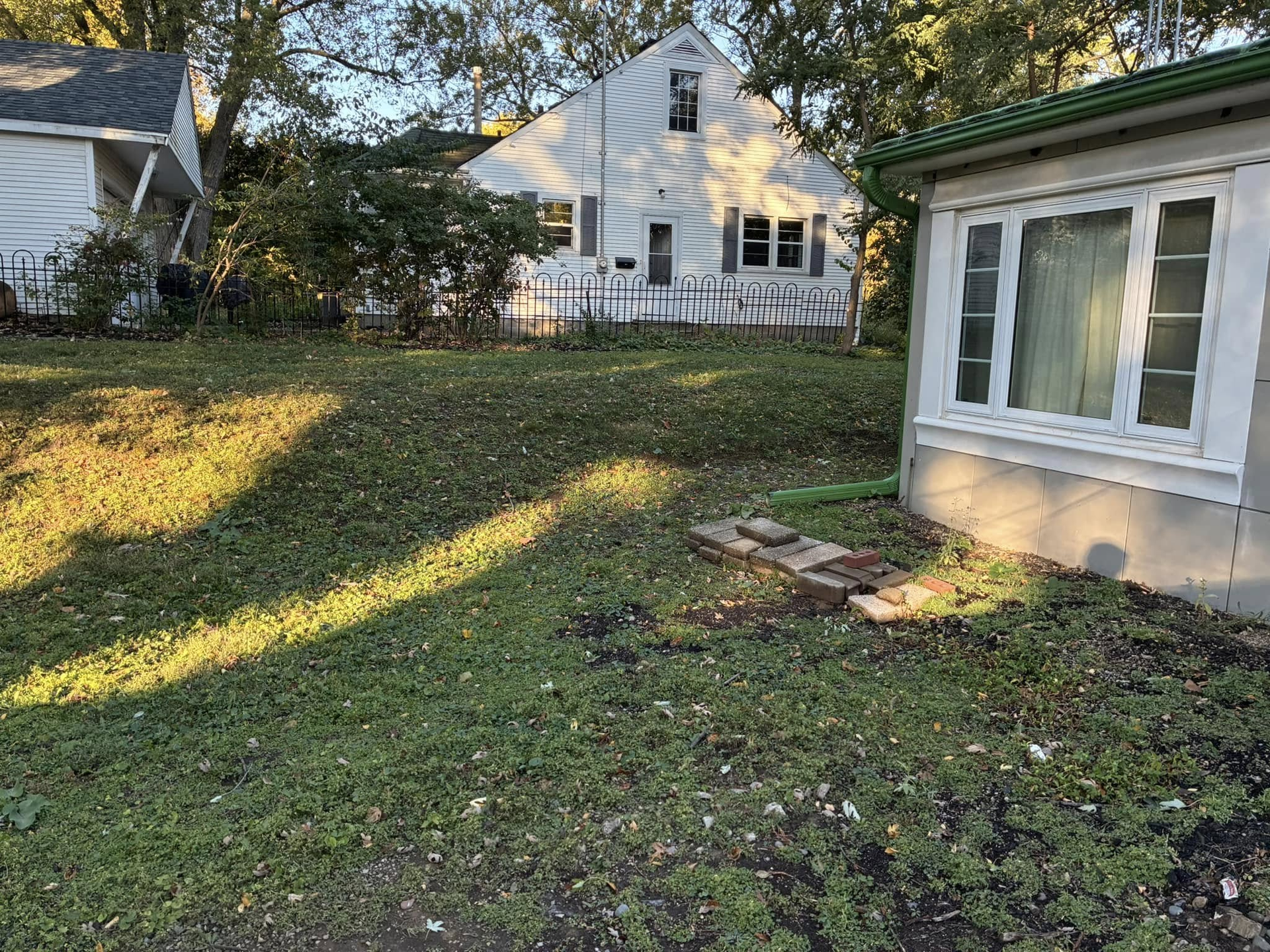 A yard with grass and overgrown foliage in front of a white house and a smaller structure.