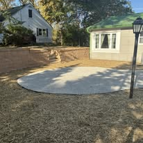 Circular concrete patio in yard, near house and retaining wall; lamp post.
