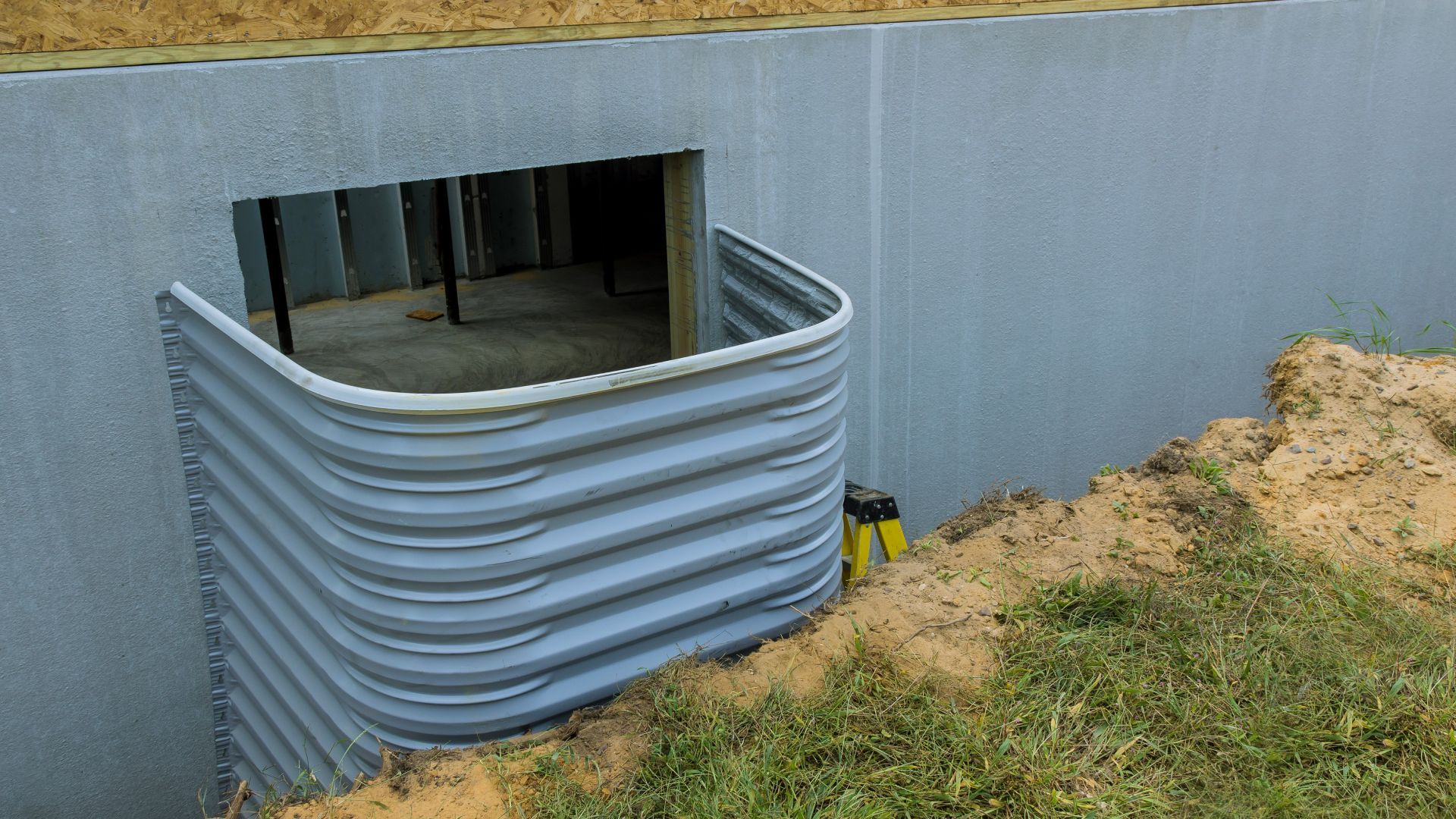 Grey corrugated metal window well installed against a concrete basement wall with exposed soil.