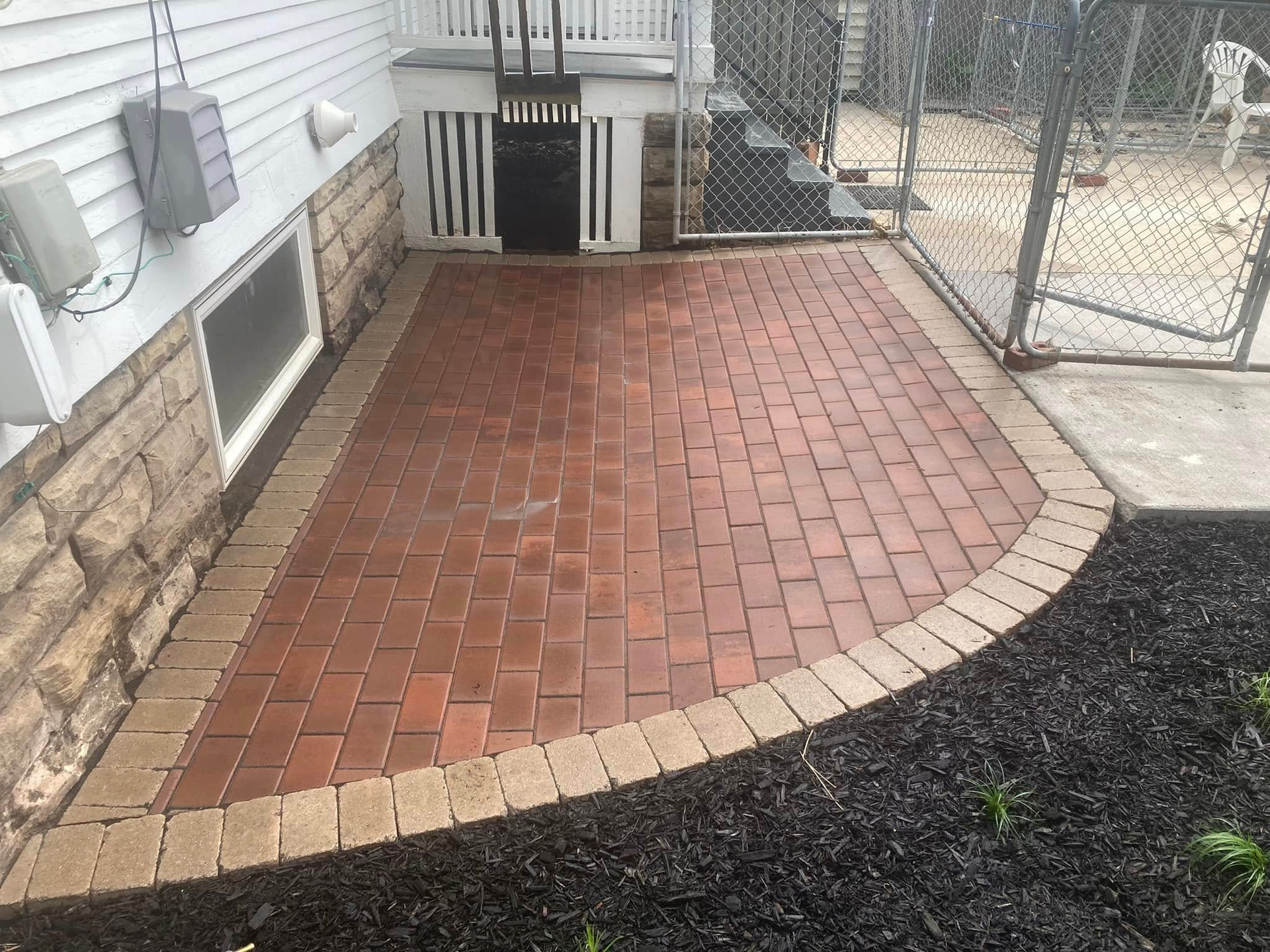 Brick patio with dark mulch border, near a house with stairs and a chain link fence.