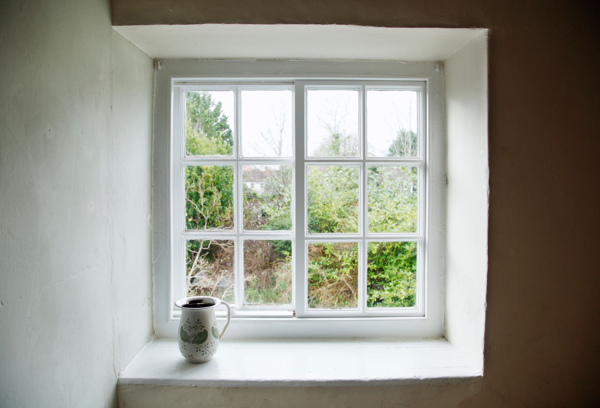 White-paned window with jug plant overlooks lush green garden.