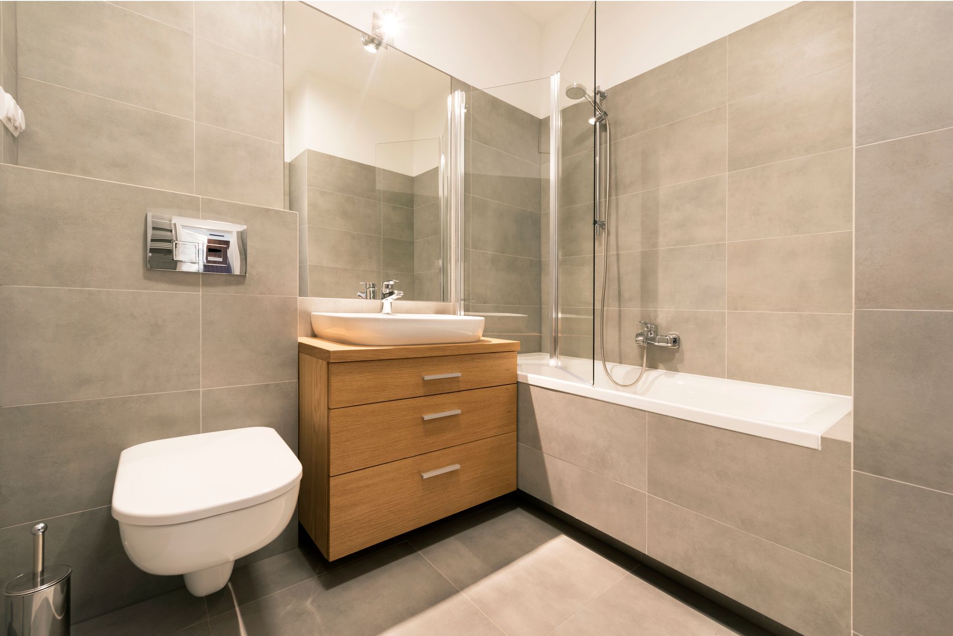Modern bathroom with gray tile walls, wood vanity, and bathtub with glass shower screen.
