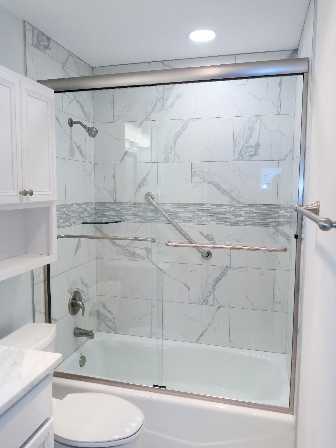 Bright bathroom with a white tub, marble-look tile, and a glass shower door. Includes a toilet, shelves, and a silver grab bar.